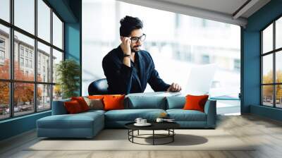 Portrait of a busy indian guy multitasking, taking notes, reading paper, surfing internet with laptop in office Wall mural